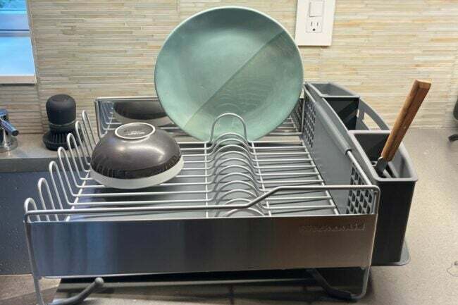 El escurridor de platos de tamaño completo de KitchenAid sobre la encimera de la cocina secando un plato azul y un tazón gris.
