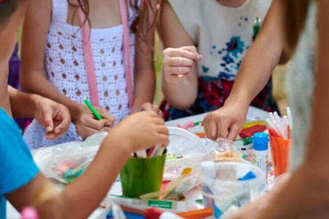 Um grupo de crianças reunidas em torno de uma mesa de artesanato com materiais de arte fazendo artesanato. 