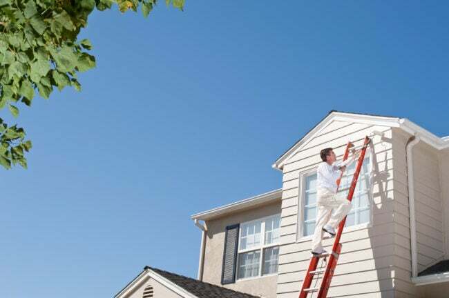 Uma pessoa sobe em uma escada extensível e pinta o segundo andar de uma casa em branco sujo.