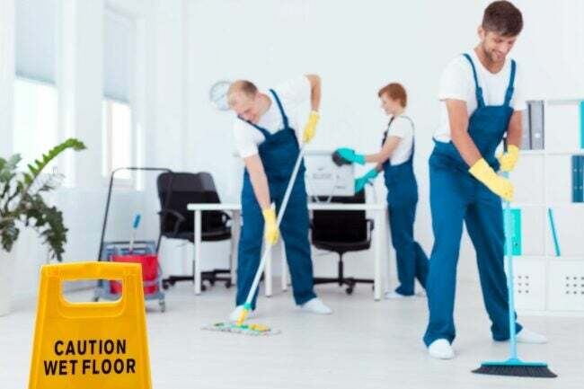 Uma equipe de faxineiros de macacão azul e luvas amarelas limpa o chão.