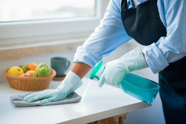 Osoba u plavoj odjeći i rukavicama za čišćenje prska otopinu na radnu površinu i briše je. 