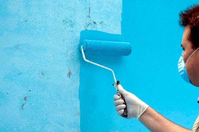 Мужчина в маске красит стену в синий цвет валиком
