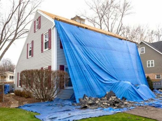 γκρι αποικιακό σπίτι καλυμμένο με μεγάλο μπλε μουσαμά με κεραμίδια στέγης στο γκαζόν ενώ η στέγη αντικαθίσταται