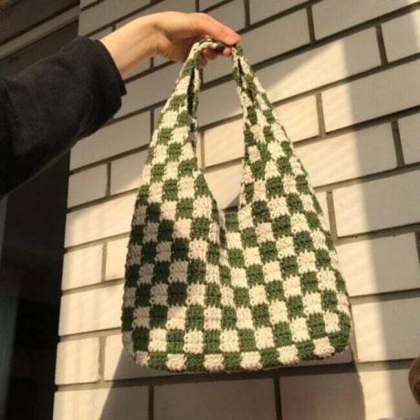 Uma bolsa de crochê xadrez verde e branco encostada em uma parede de tijolos claros