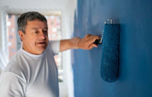 чоловік у білій футболці за допомогою валика наносить другий шар темно-синьої фарби на стіну