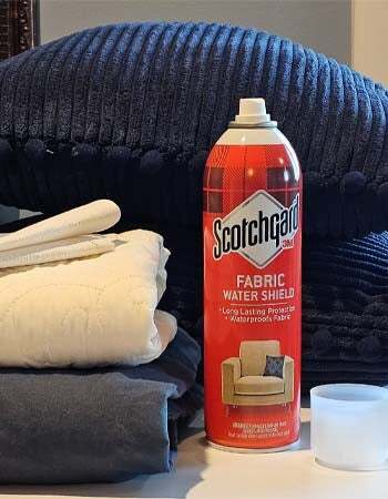 O protetor de tecido Scotchgard em uma mesa ao lado de uma variedade de tecidos, incluindo lençóis e estofados.
