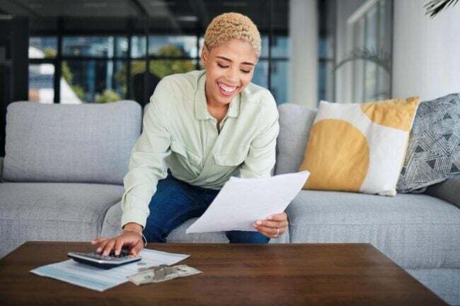 Moteris šypsosi sėdėdama ant pilkos sofos, žiūrėdama į dokumentą ir spausdindama skaičiuotuvu ant rudo kavos staliuko. 