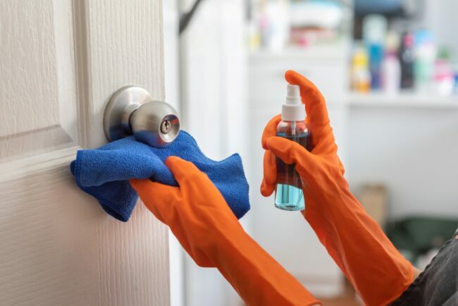 Persona che indossa guanti di gomma arancioni che disinfettano la maniglia della porta utilizzando spray e un panno blu.