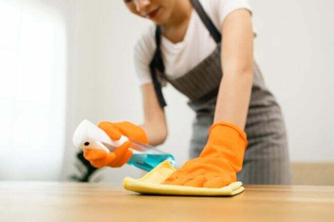 Eine Person in orangefarbenen Handschuhen sprüht eine Reinigungslösung auf einen Tisch und wischt sie mit einem gelben Tuch ab.