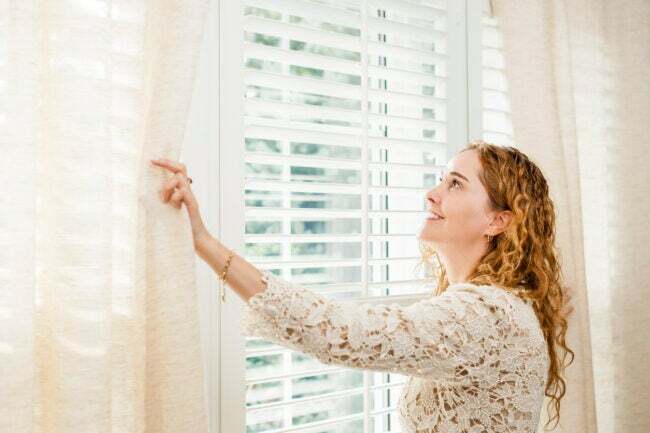 mulher na janela iluminada pelo sol sorrindo e abrindo as persianas