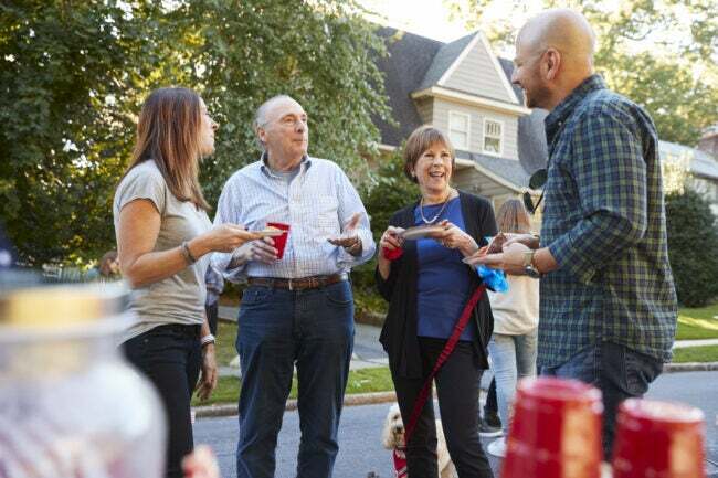 Vizinhos de meia-idade e idosos conversando em uma festa do quarteirão segurando copos de plástico vermelhos na frente de uma casa grande
