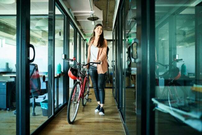 Genç iş kadını bisikletini itiyor ve ofiste kabinlerin yanından geçiyor.