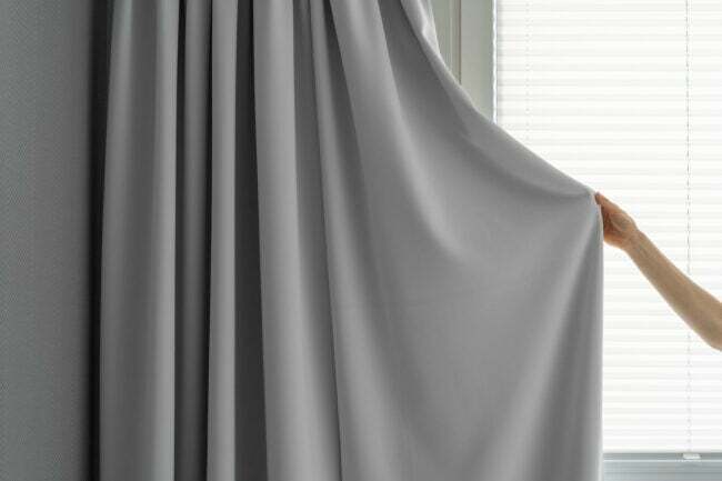 rajattu kuva naisesta pitele harmaata kangasverhoa ja sulje ikkuna auringonvalolta suojaamiseksi olohuoneessa modernilla tyylillä, yksityisyyden ja mukavuuden konseptilla