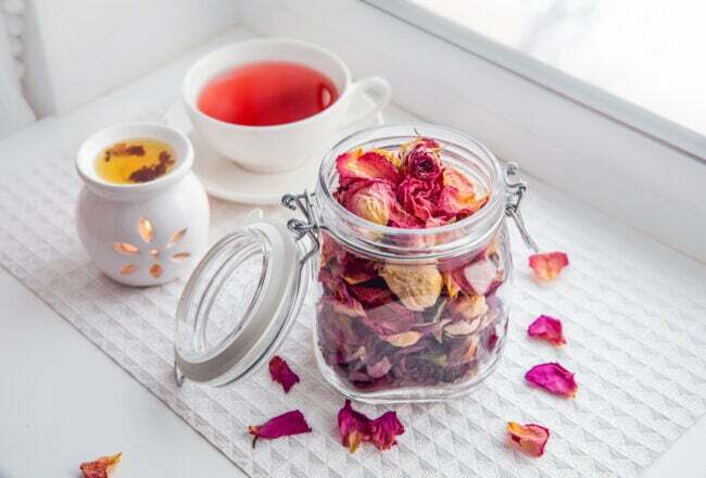 En öppen burk med torkat rosenbladpotpurri bredvid en liten eterisk oljevärmare och en kopp te