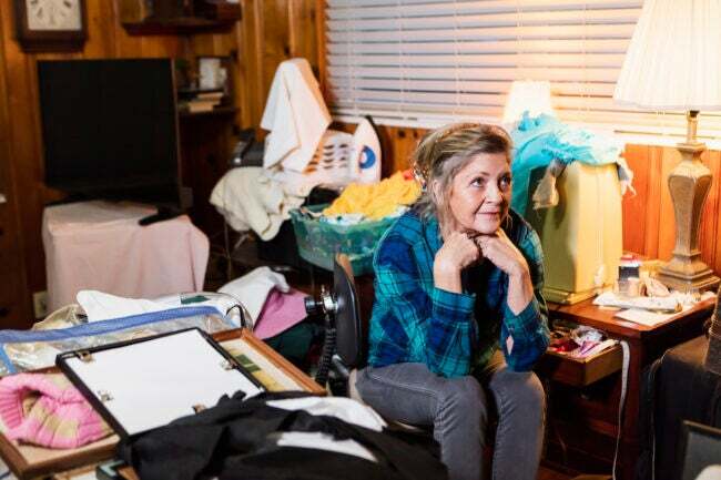 O femeie în vârstă de 60 de ani acasă, stând într-o cameră dezordonată, aglomerată, privind în altă parte cu o expresie serioasă.