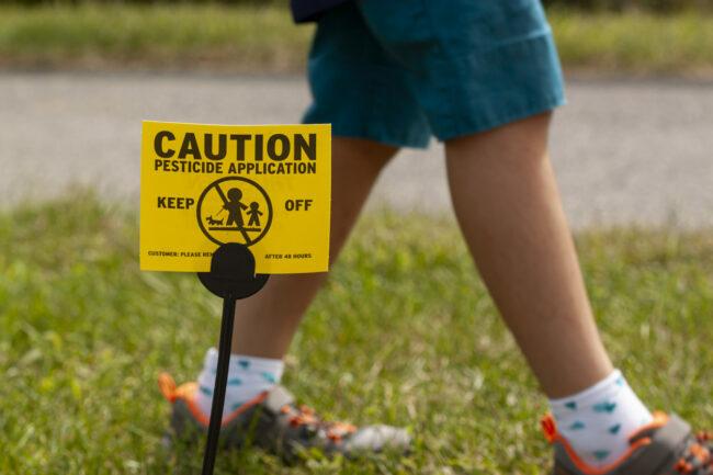 Clarksburg, MD, ZDA 09. 12. 2020: Rumen znak na dvorišču, ki otroke in hišne ljubljenčke opozarja na nedavno škropljenje s pesticidi in jim svetuje, naj se izogibajo. Ne glede na to se otrok igra. Uporaba pesticidov je velika skrb.