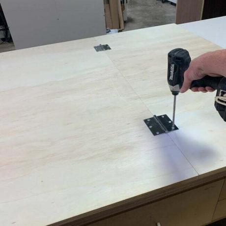 Seorang tukang kayu memasang engsel pada kayu lapis untuk membuat bagian atas meja kerja.