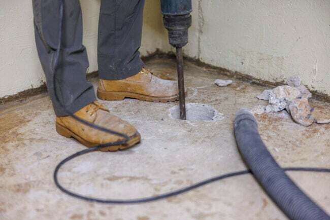 Instalace protiradonového systému. Dělník vrtá díru do podlahy sklepa, aby zlepšil dům.