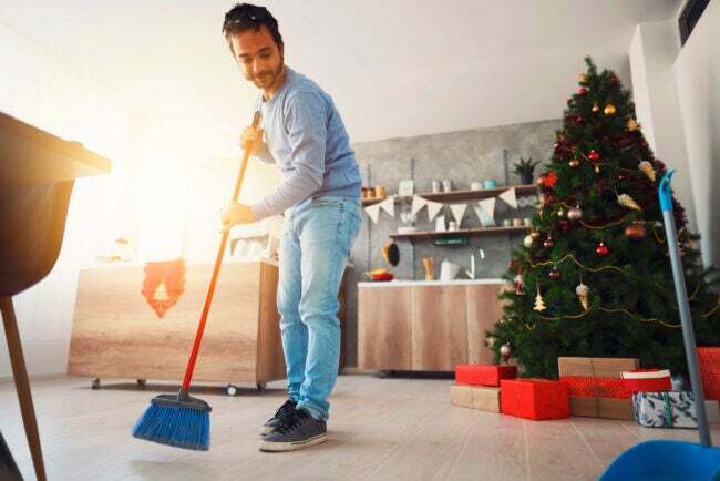 Muž, ktorý zametá podlahu metlou a lopatkou na prach vo svojom dome, s vianočným stromčekom za ním.