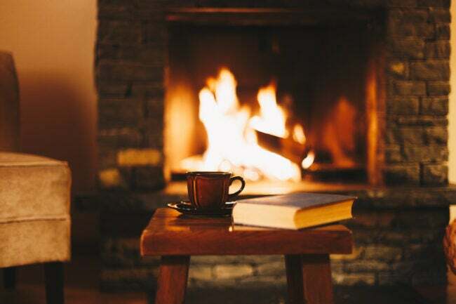 scaun cu cană și carte în fața unei flăcări calde deschise a șemineului în camera de zi confortabilă
