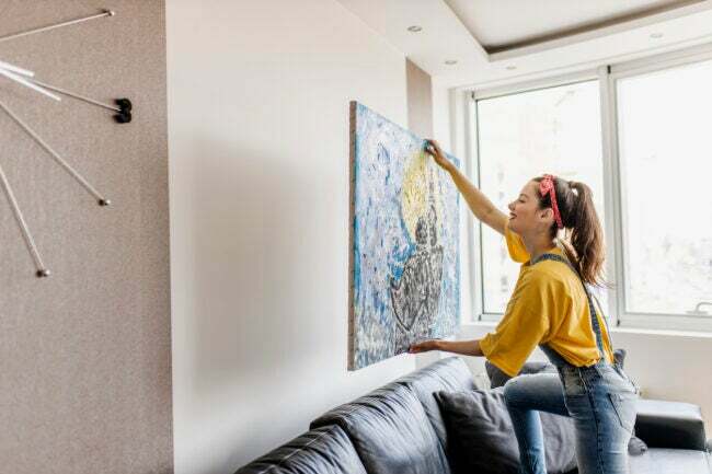 युवा महिला दीवार पर कला चित्र टांग रही है और लिविंग रूम को सजा रही है