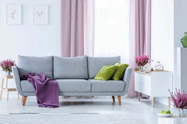 Серый диван с фиолетовым пледом и зелеными подушками перед розовыми шторами в гостиной