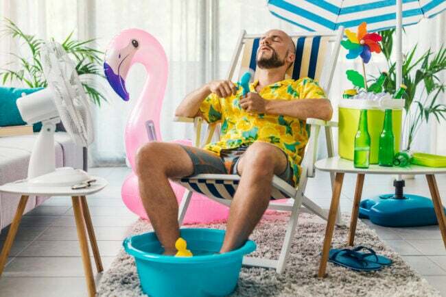 Homem se refrescando com ventiladores elétricos e sentado em uma espreguiçadeira dentro de casa, com flamingo inflável