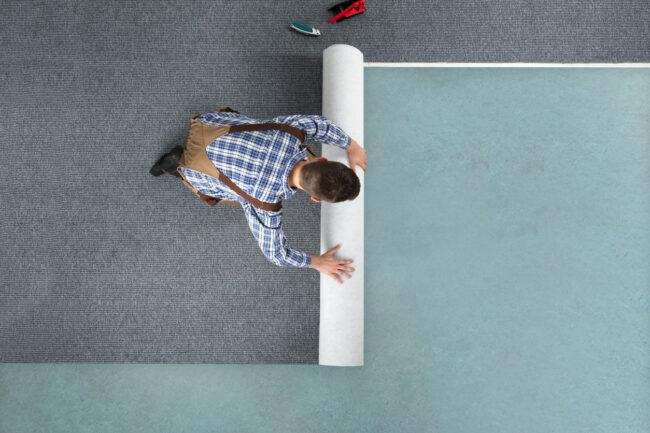מבט בזווית גבוהה לעובד צעיר באוברול שטיח מתגלגל על ​​הרצפה בבית