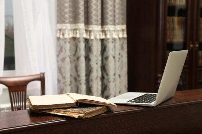 Na drevenej rímse sedí otvorený laptop spolu s historickými knihami a starožitným nábytkom.