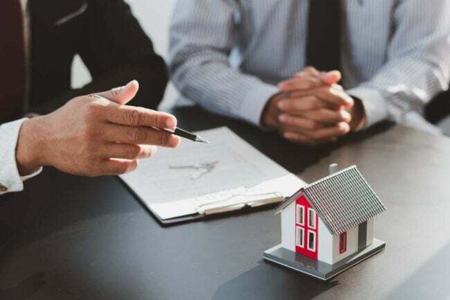 Una pequeña casa modelo aparece sobre una mesa junto a dos pares de manos que gesticulan sobre un documento.