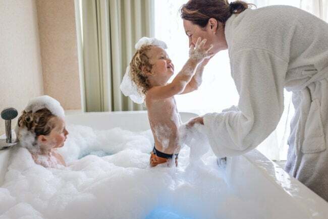 امرأة في الأبيض رداء حمام تميل على فقاعة حمام وتلعب مع صبي بينما صبي آخر ينظر على