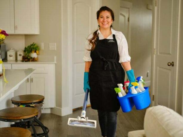 Uma mulher sorridente com um avental preto segura um transportador azul com produtos de limpeza e uma ferramenta de limpeza e está em uma cozinha.