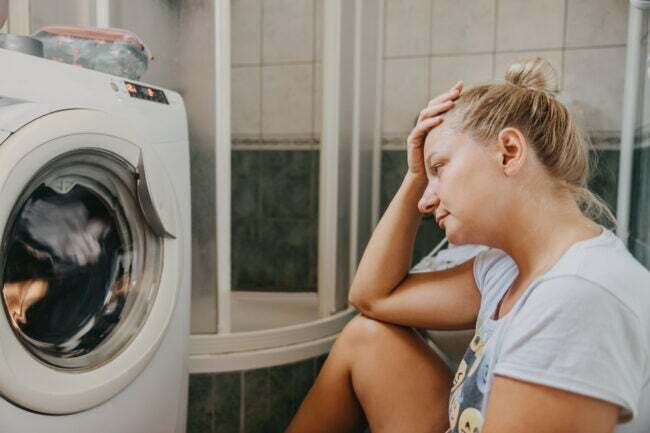 Засмучена жінка сидить у пральні й дивиться на пральну машину