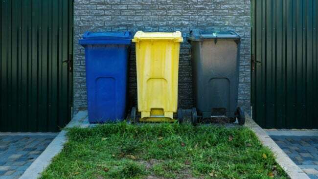 Синие желтые и серые пластиковые мусорные баки стоят в ряд на заднем дворе
