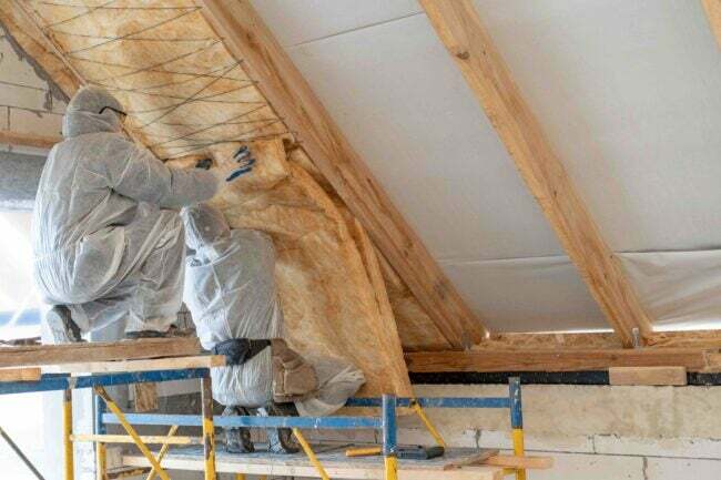 Achteraanzicht van twee professionele arbeiders in overalls die samenwerken met steenwolisolatie, in een nieuw huis in aanbouw staan, warmtemateriaal aan plafond en muur bevestigen met kopieerruimte