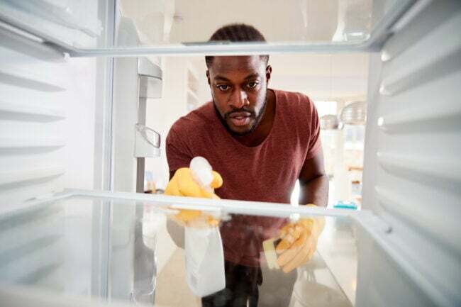 Vue depuis l'intérieur d'un réfrigérateur vide alors qu'un homme portant des gants en caoutchouc nettoie les étagères.