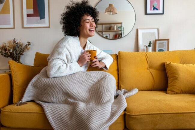 kvinna skrattar sitter på soffan täckt av filt och håller en mugg