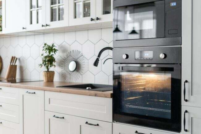 Šiuolaikinio juodos orkaitės virtuvės interjero dizaino vaizdas iš šono. Stilingas balto medžio baldo paviršius su buitine technika ir dekoru. Jauki vieta maisto gaminimui. Kepimo procesas