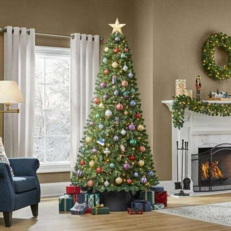 Home Accents Holiday Prelit LED šventinė pušis papuošta ornamentais ir žvaigždės viršūne su dovanomis apačioje ir dekoruotu židiniu fone.