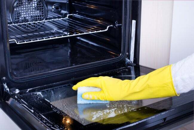 Lähikuva naisen kädestä keltaisilla suojakäsineillä puhdistamassa uunin luukkua