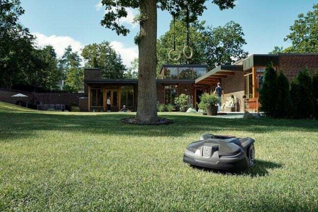 Mesin pemotong rumput robotik Husqvarna di halaman hijau besar dengan latar belakang rumah yang luas.