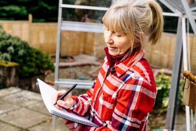 Kvinde iført en rød og hvid skjorte og skriver haveplaner i en notesbog udendørs.