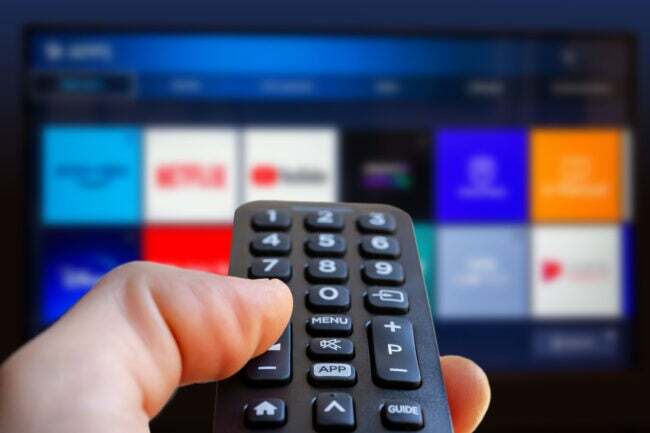 एक आदमी अपने हाथ में स्मार्ट टीवी का रिमोट कंट्रोल पकड़े हुए है और बैकग्राउंड में टीवी स्क्रीन है।
