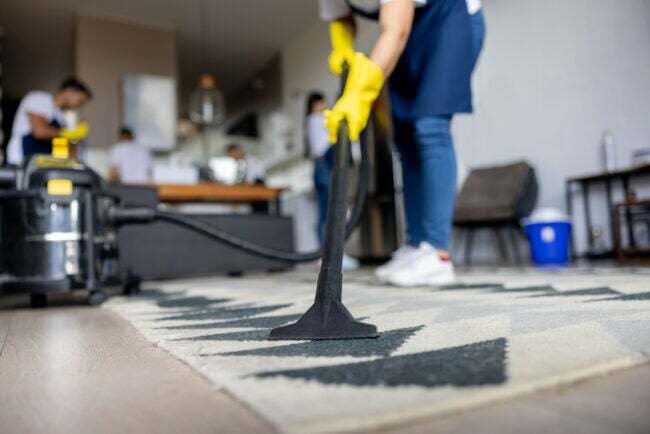 Una persona pulisce un tappeto mentre una squadra di addetti alle pulizie lavora in cucina. 