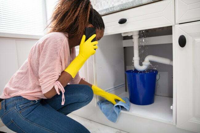 en-kvinde-i-lyserød-skjorte-og-jeans-bruger-gule-gummi-handsker-mens-i-foretager-et-telefonopkald-og-kigger-på-en-lækage-under-vasken-fanget- ved en spand