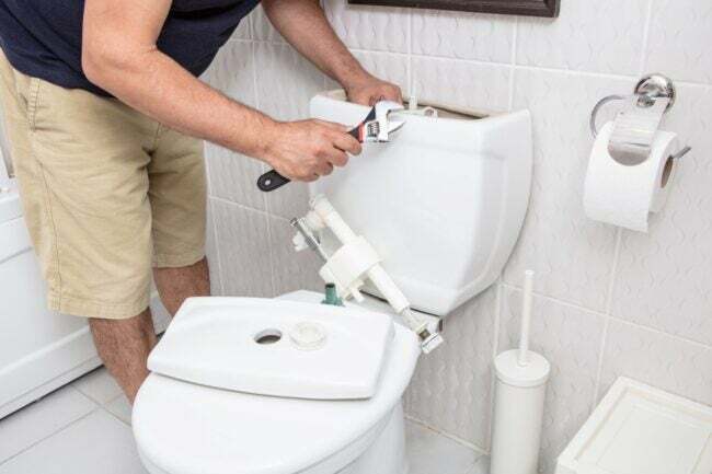 Mājas īpašnieks demonstrē, kā nomainīt tualetes uzpildes vārstu ar regulējamu uzgriežņu atslēgu