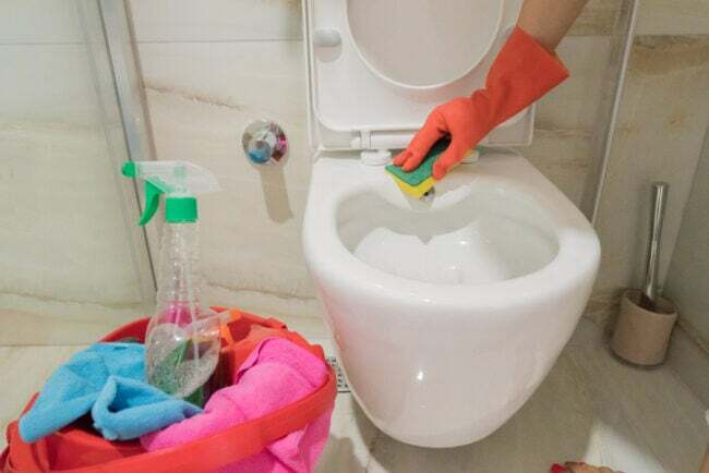 hand-in-organge-handske-använder-svamp-för-att-städa-toalett-med-rosa-korg-med-städmaterial-i närheten-i-beige-badrum