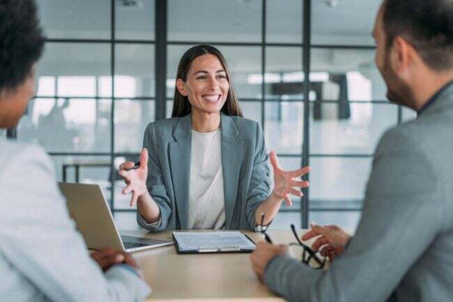 Uma mulher de blazer cinza gesticula enquanto fala e sorri para duas pessoas sentadas à sua frente em um escritório.