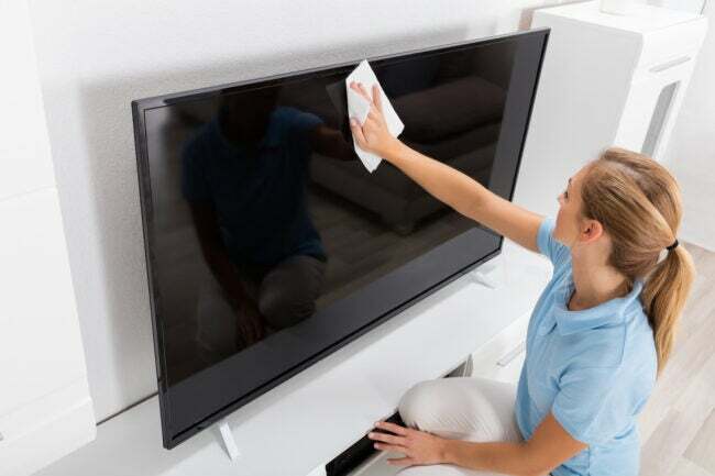 Fiatal nő tisztító TV képernyőt egy fehér nappaliban