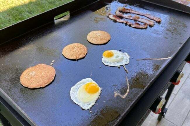 Três panquecas, dois ovos e três tiras de bacon cozinhando em uma frigideira.